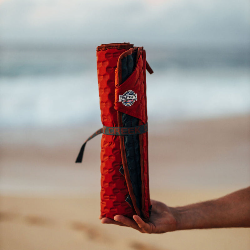 Load image into Gallery viewer, Crazy Creek Hex Chair rolled up and secured with strap - Man holding it in front of a beach
