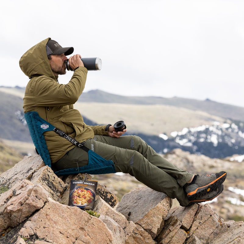 Load image into Gallery viewer, Man drinking water while sitting in a blue crazy creek chair while sitting on the top of a mountain