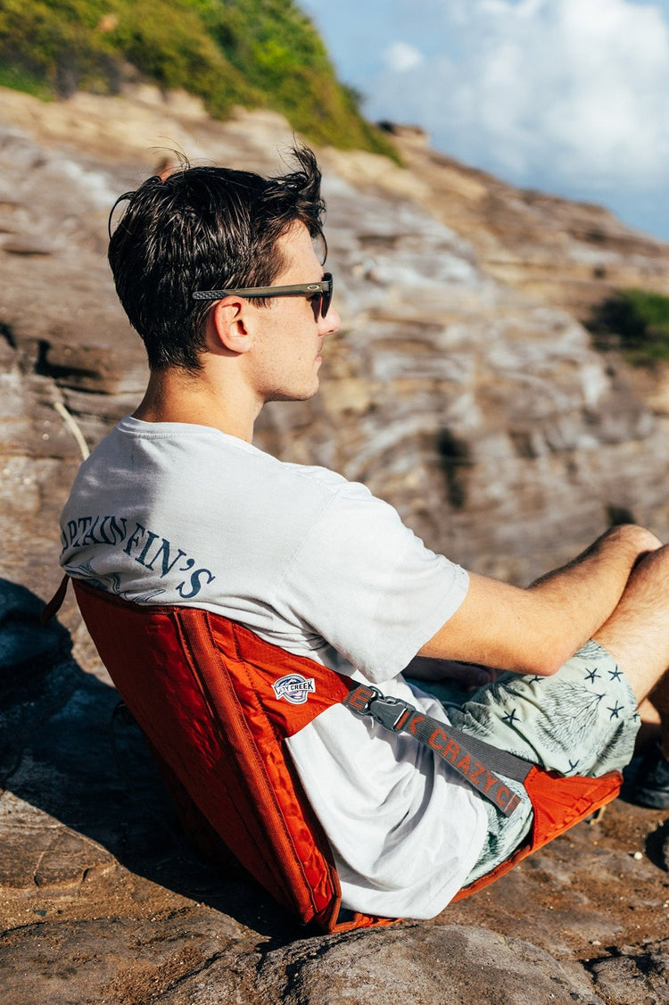 Man sitting on a red Crazy Creek HEX 2.0 Chair, enjoying portable comfort in an outdoor setting.