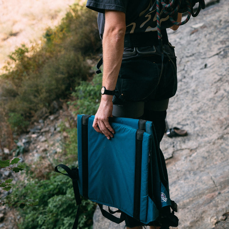 Man carrying a blue Crazy Creek The Chair along a scenic trail.