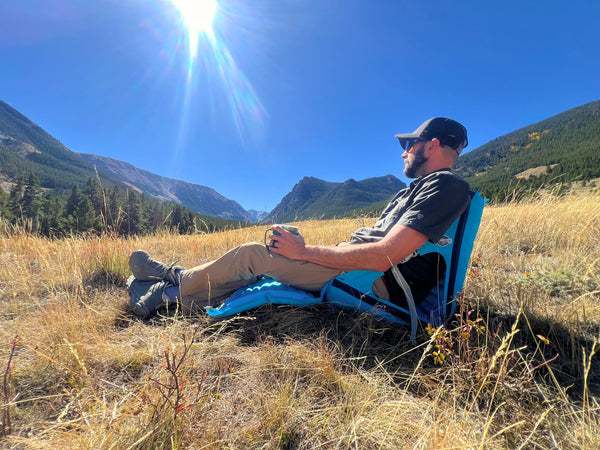 Man sitting on a blue camping chair in a mountainous landscape with mountains and trees in the background.