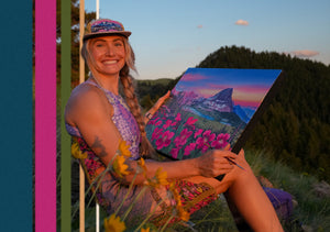 Rachel Pohl sitting outdoors with a painting of a mountain landscape while sitting in her limited edition crazy creek chair. 