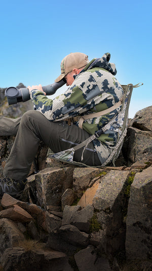 Hunter sitting in a camo crazy creek chair holding a monocular