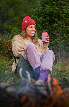 Woman holding a drink sitting in a crazy creek chair in front of a bonfire