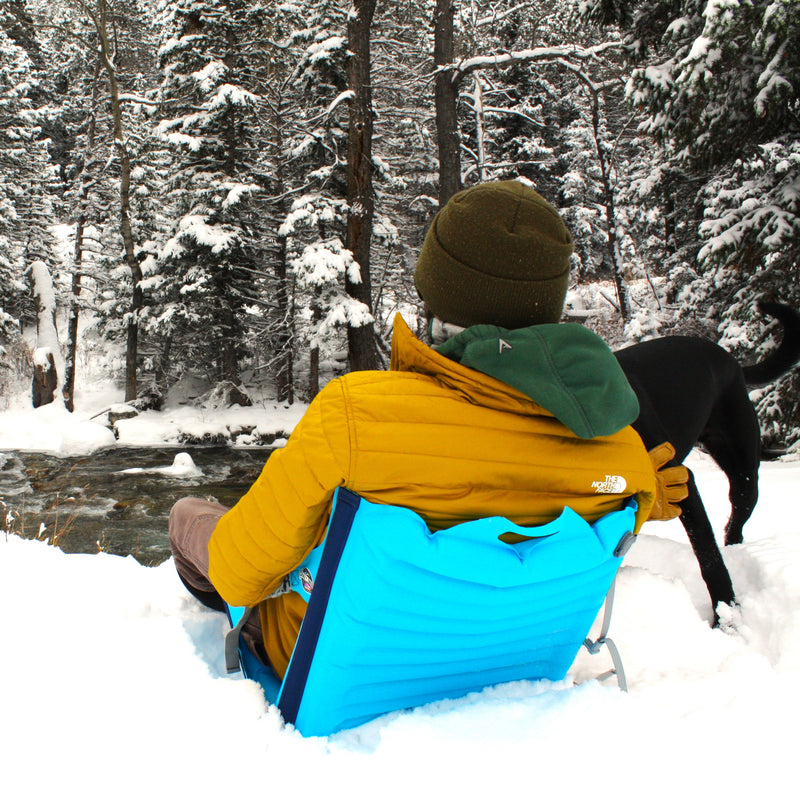 Load image into Gallery viewer, Person sitting in a blue inflatable chair with his dog next to a winter river in the snow
