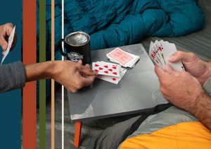 Two people playing cards on a small crazy Creek folding table with a coffee mug and colorful vertical stripes in the background.