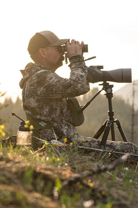 Man sitting on a Crazy Creek HEX 2.0 Original Chair in Olive/Slate, dressed in camouflage and using binoculars in a natural outdoor environment.