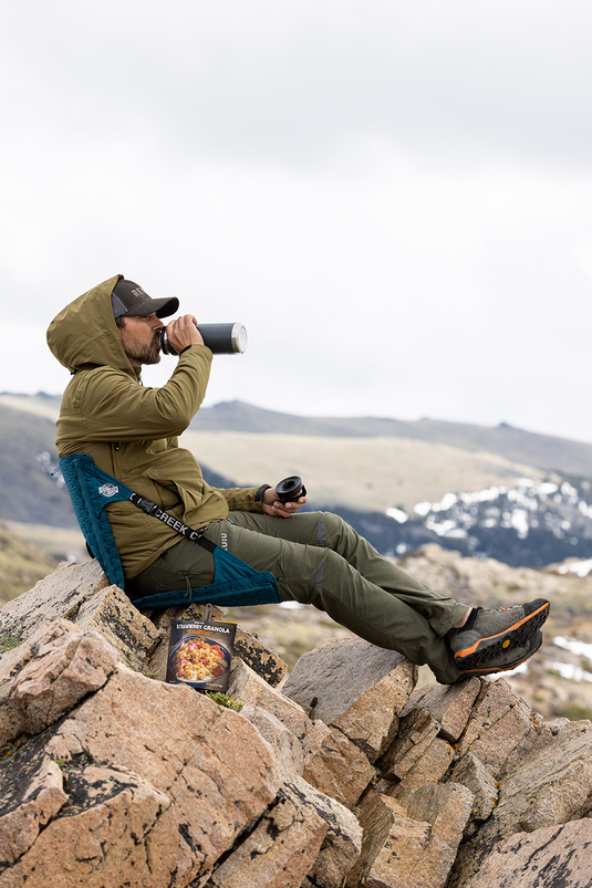 Man sitting in a Crazy Creek HEX 2.0 Original Chair, sipping from a bottle while enjoying a scenic hillside view.