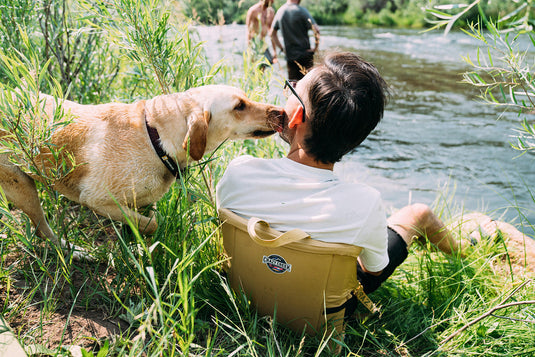 Man relaxing by a river with his dog on a Crazy Creek LongBack Chair, enjoying the outdoors together.