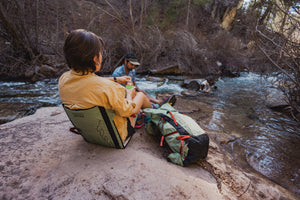 Woman and Man relaxing on the bank of a river with their Osprey x Crazy Creek Talon backpack and Chair