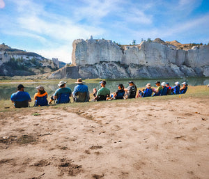 people lined up in their crazy creek chairs on the bank of a lake eating lunch and talking