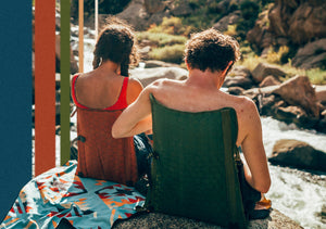 a couple sitting in green and copper chairs in front of a babbling brook in the mountains