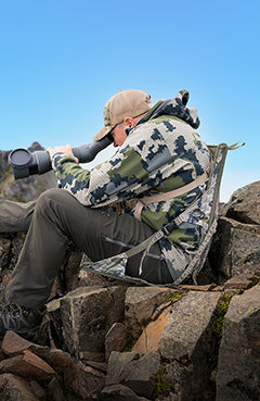 Hunter sitting in a camo crazy creek chair holding a monocular