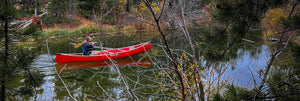 A wide shot of a man paddling a red canoe down a river while sitting in a crazy creek chair