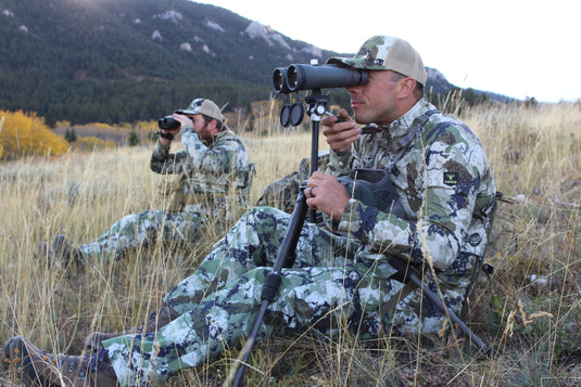 Man sitting on a Crazy Creek King’s Camo Original Chair, dressed in camouflage and using binoculars in a natural outdoor setting.