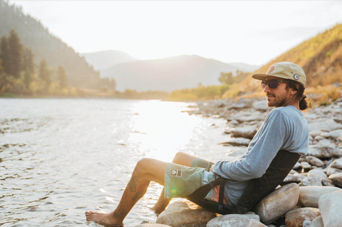 Man relaxing by a river on a Crazy Creek LongBack Chair, enjoying the scenic outdoor setting.