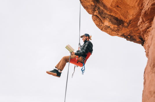 Man tethered to a rope, suspended below a rocky overhang, sitting in a Crazy Creek LongBack Chair and reading a book.