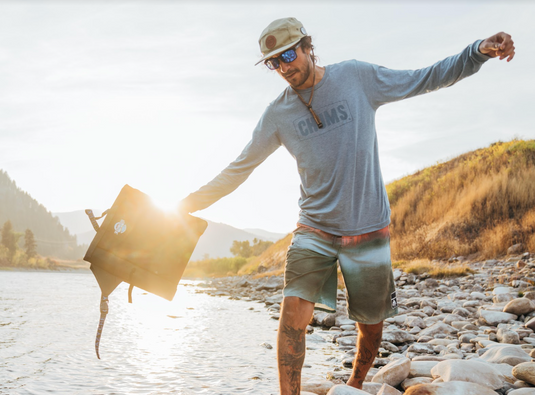 Man carrying a Crazy Creek Original Chair along the rocky shore of a lake, heading to his next adventure.