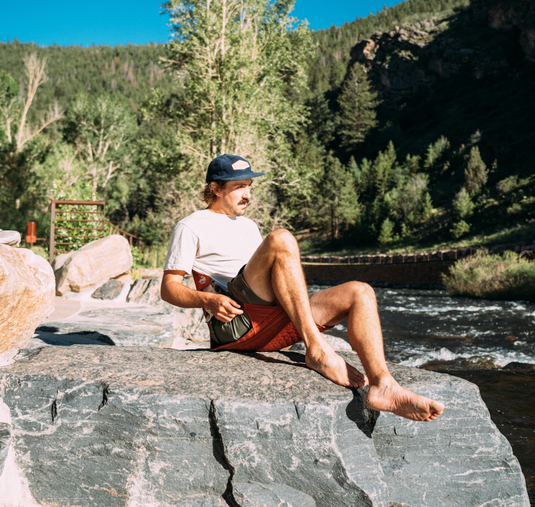 Man reclining in a Crazy Creek HEX 2.0 LongBack Chair by a river, enjoying a relaxing outdoor setting.