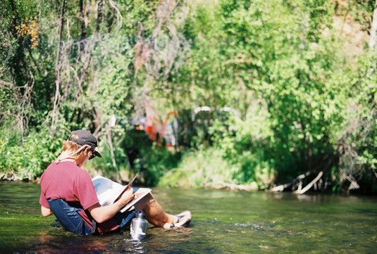 Man relaxing in a shallow river, reading a book while seated in a Crazy Creek Original Chair.